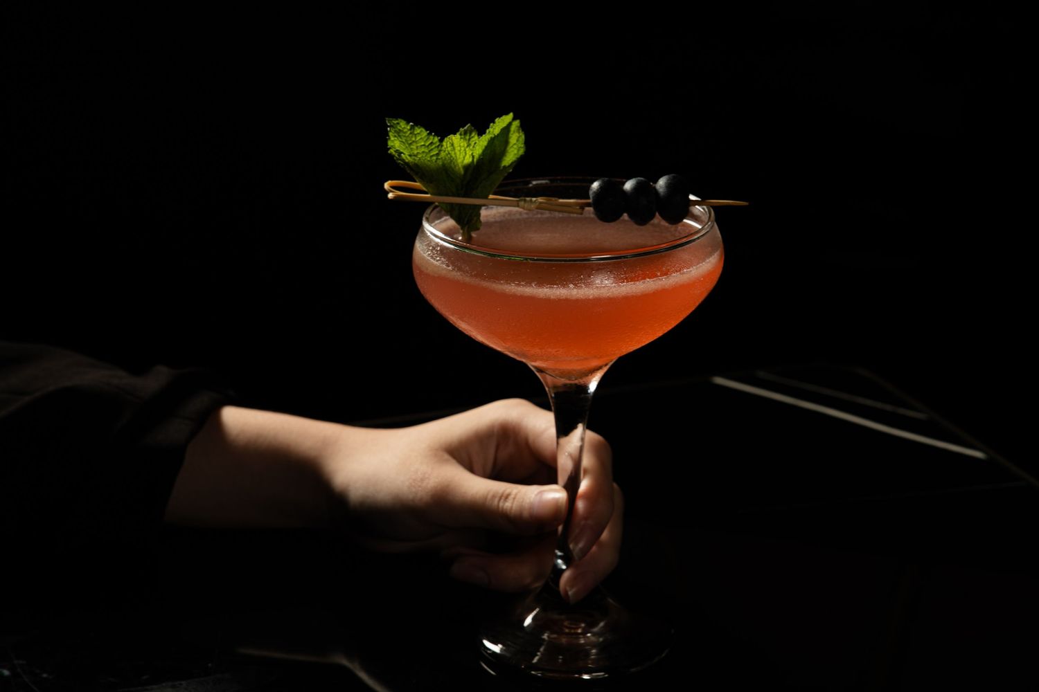 Hand holding elegant cocktail glass with red drink garnished with fresh mint leaves against dark background.