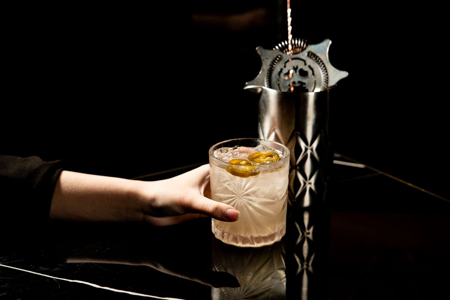A hand reaches for an artisanal cocktail in a crystal glass next to an ornate silver bar tap against a dark background.