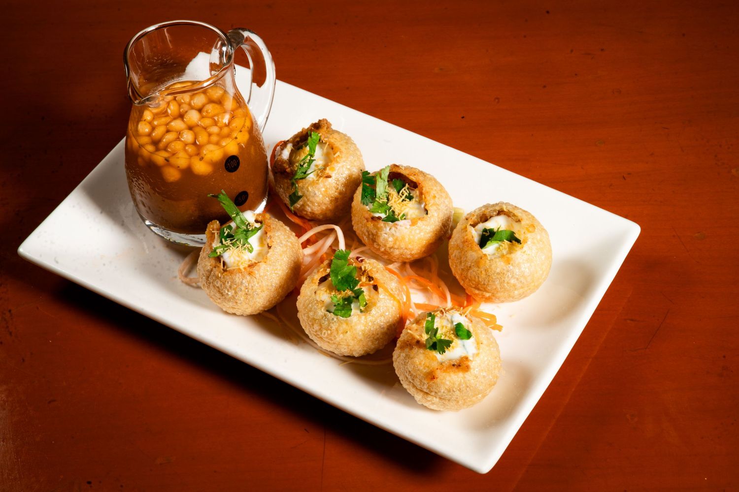 Five pani puri appetizers served on a white rectangular plate with orange dipping sauce in a glass on brown table.