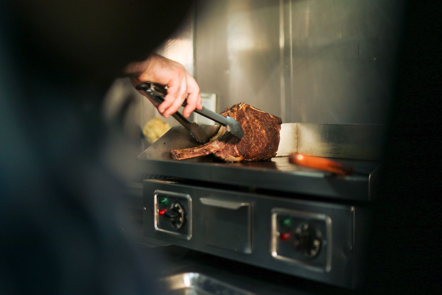 Hand carving slices from a roasted meat on a dark commercial stove surface in a dimly lit kitchen.