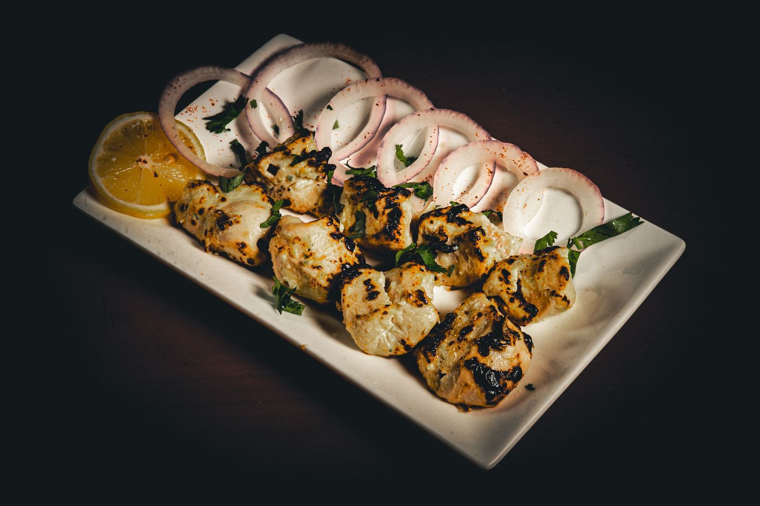 A plate of grilled chicken skewers with mushrooms and a dipping sauce served on a white rectangular platter.
