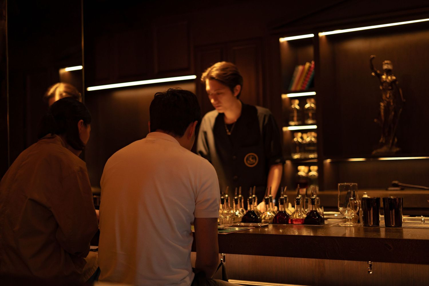 A bartender serves customers at a dimly lit upscale bar with glowing glassware displays in the background.