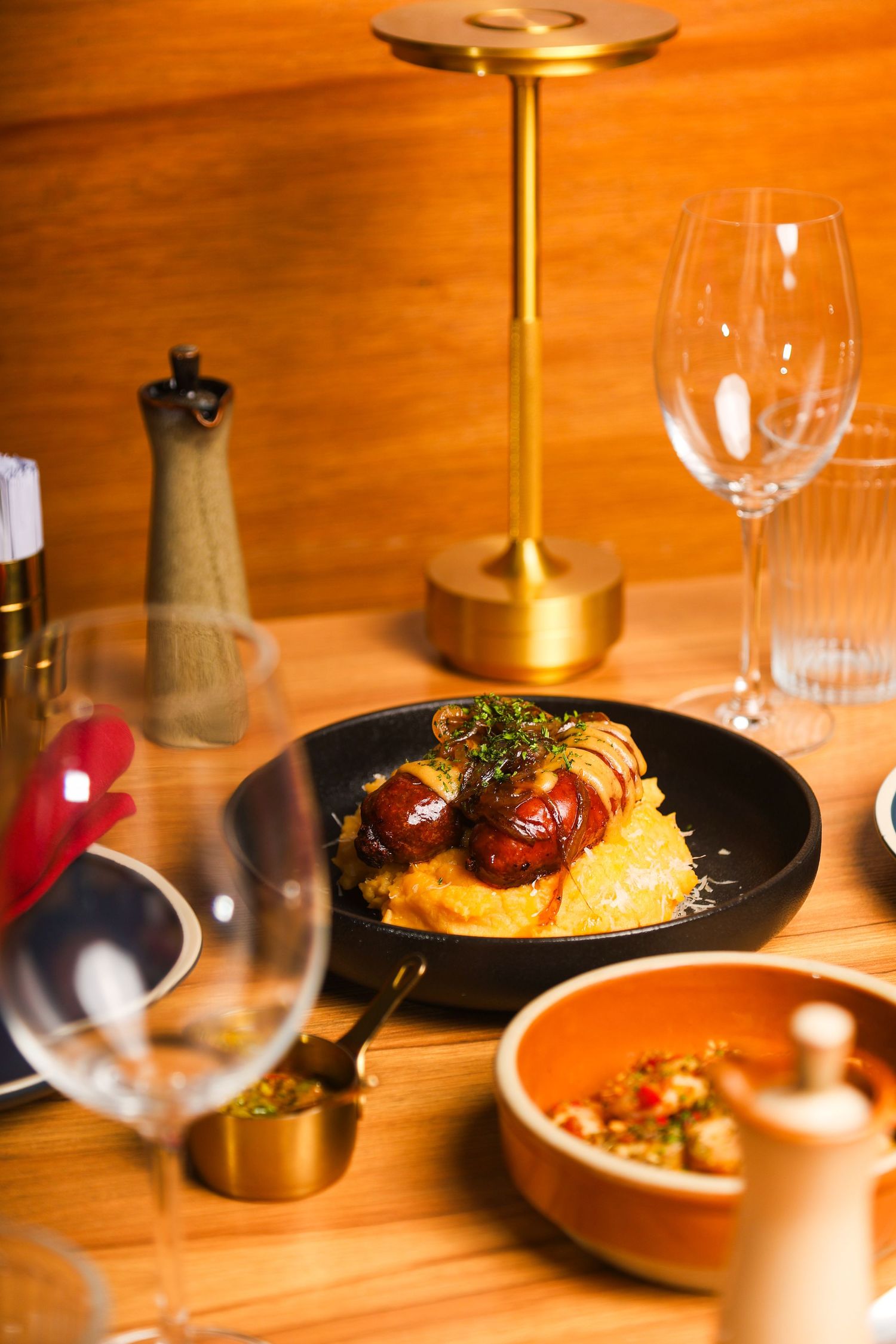 Elegant dinner setting with polenta and tomatoes on black plate alongside wine glasses and ambient lighting.