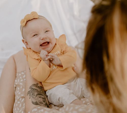 Baby in yellow outfit smiling while sitting on white bedding.