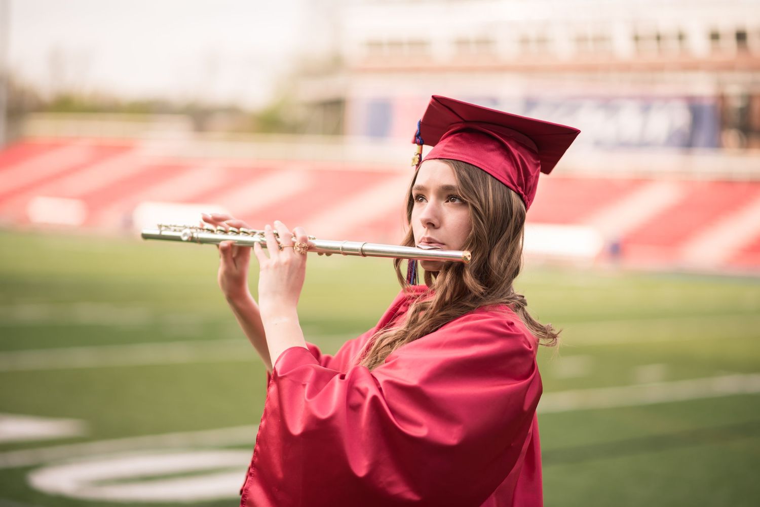 senior portrait of a graduating student in her red cap and gown, standing on the high school football field and playing her flute