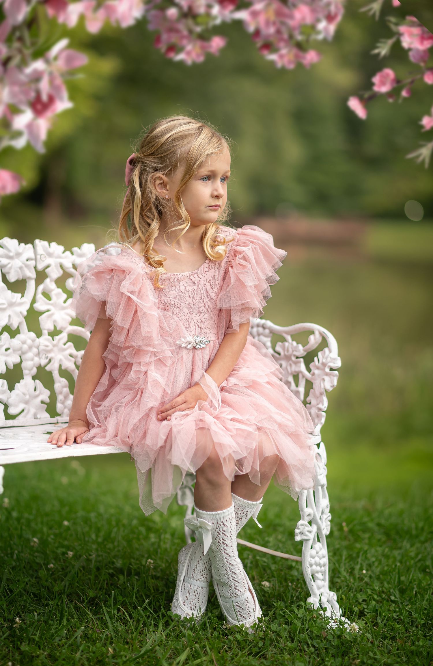 a little girl in a fluffy pink dress sits on a white bench surrounded by pink flowers and green grass