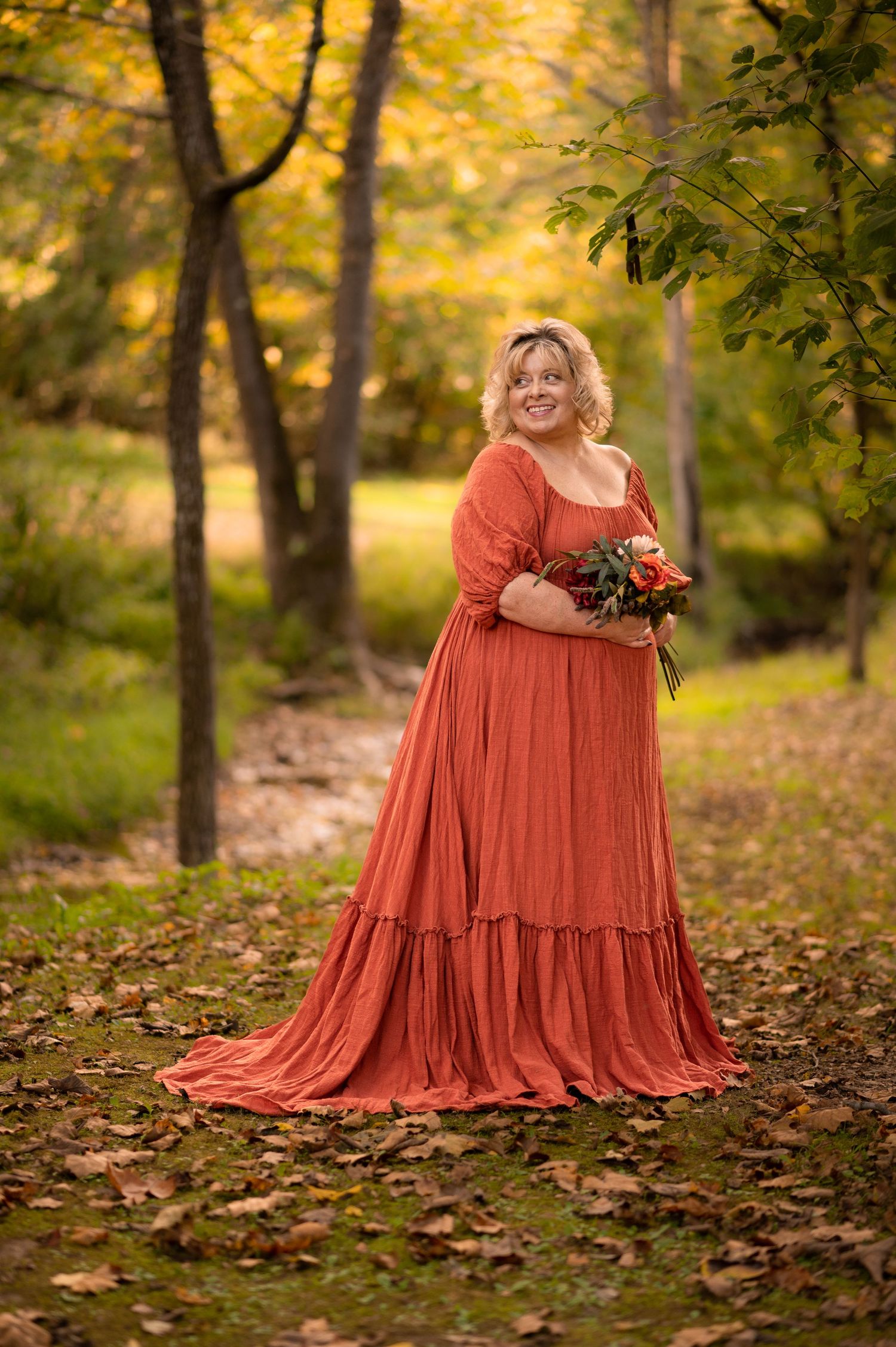 a woman in a floor length orange dress holds a bouquet of flowers and smiles, standing in the woods with sunlight behind her