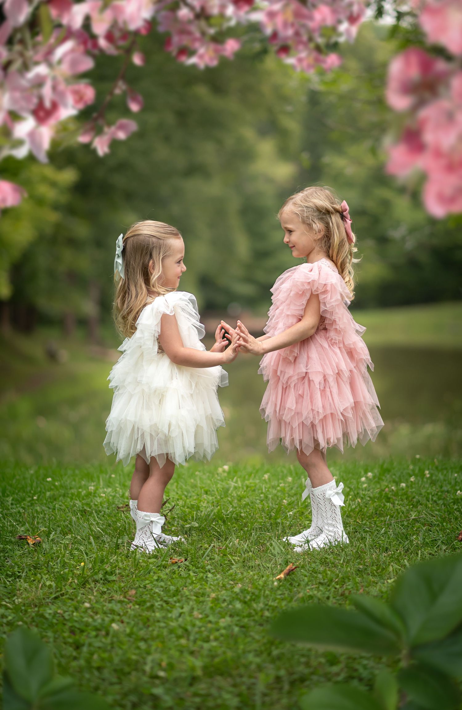 two little girls in matching pink and white fluffy tulle dresses and white tights stand in a green meadow with pink flowers overhead