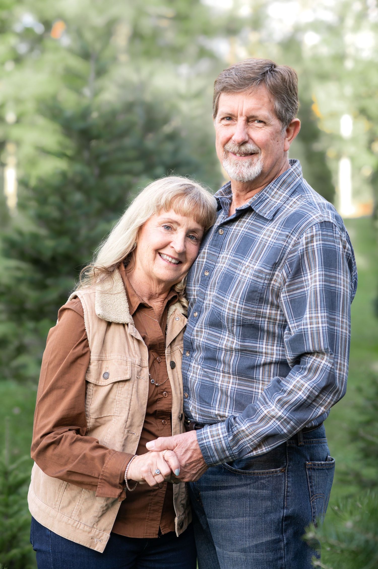 an older couple pose for a portrait, holding hands and smiling, with Christmas trees in the background
