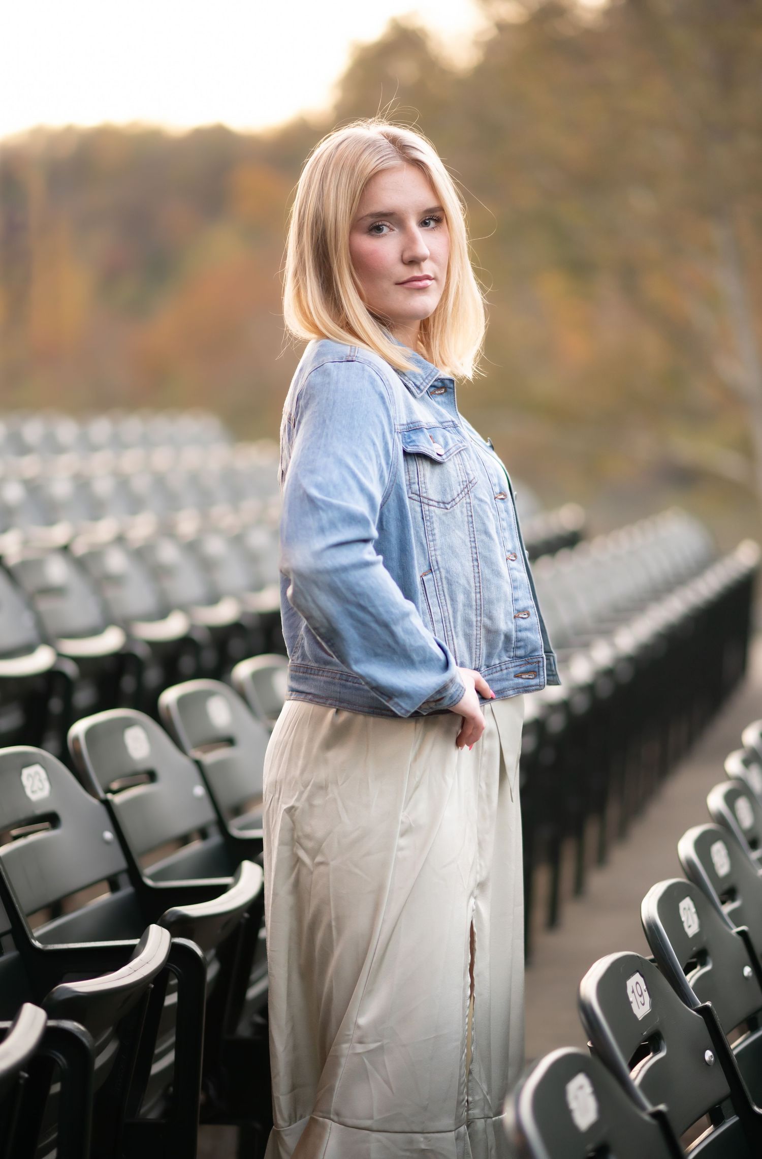 a lovely blonde high school graduate poses for a senior portrait among rows of stadium seating, with autumn trees in the background.