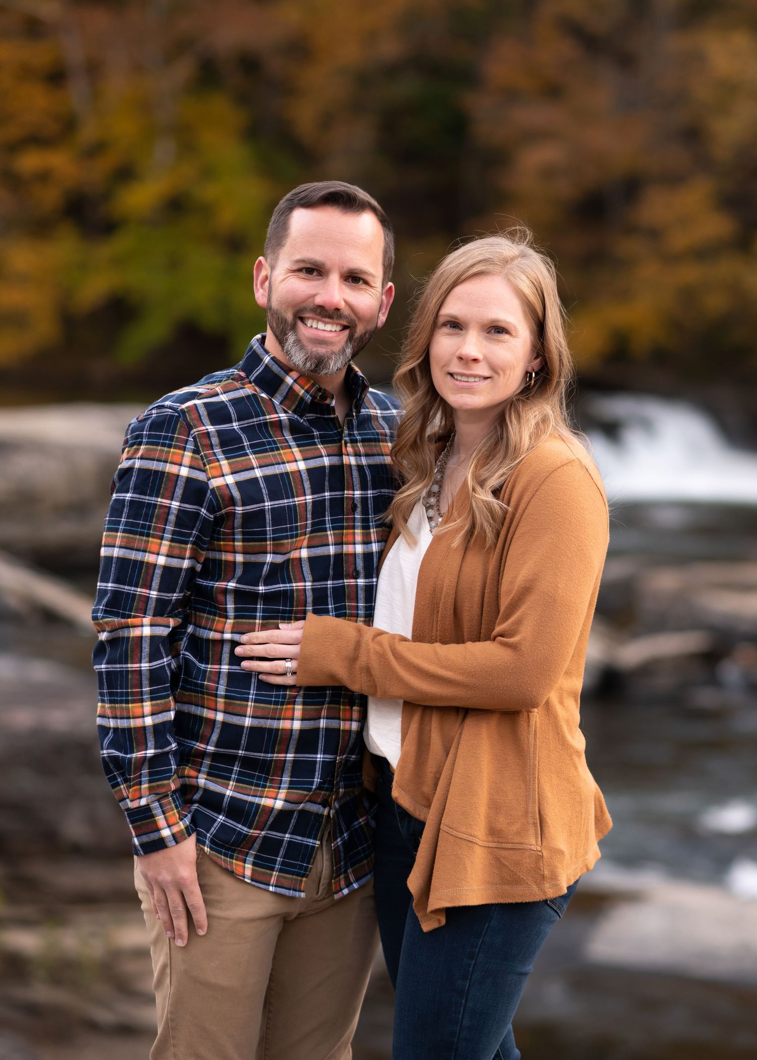 a young husband and wife stand together and pose for a portrait in front of a waterfalls on the edge of a riverbank in Fairmont, WV