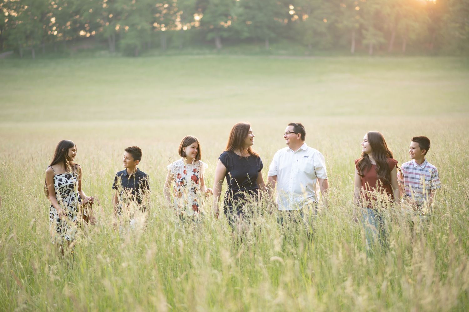 a family of six walks through a sunlit field of tall grasses, holding hands and laughing