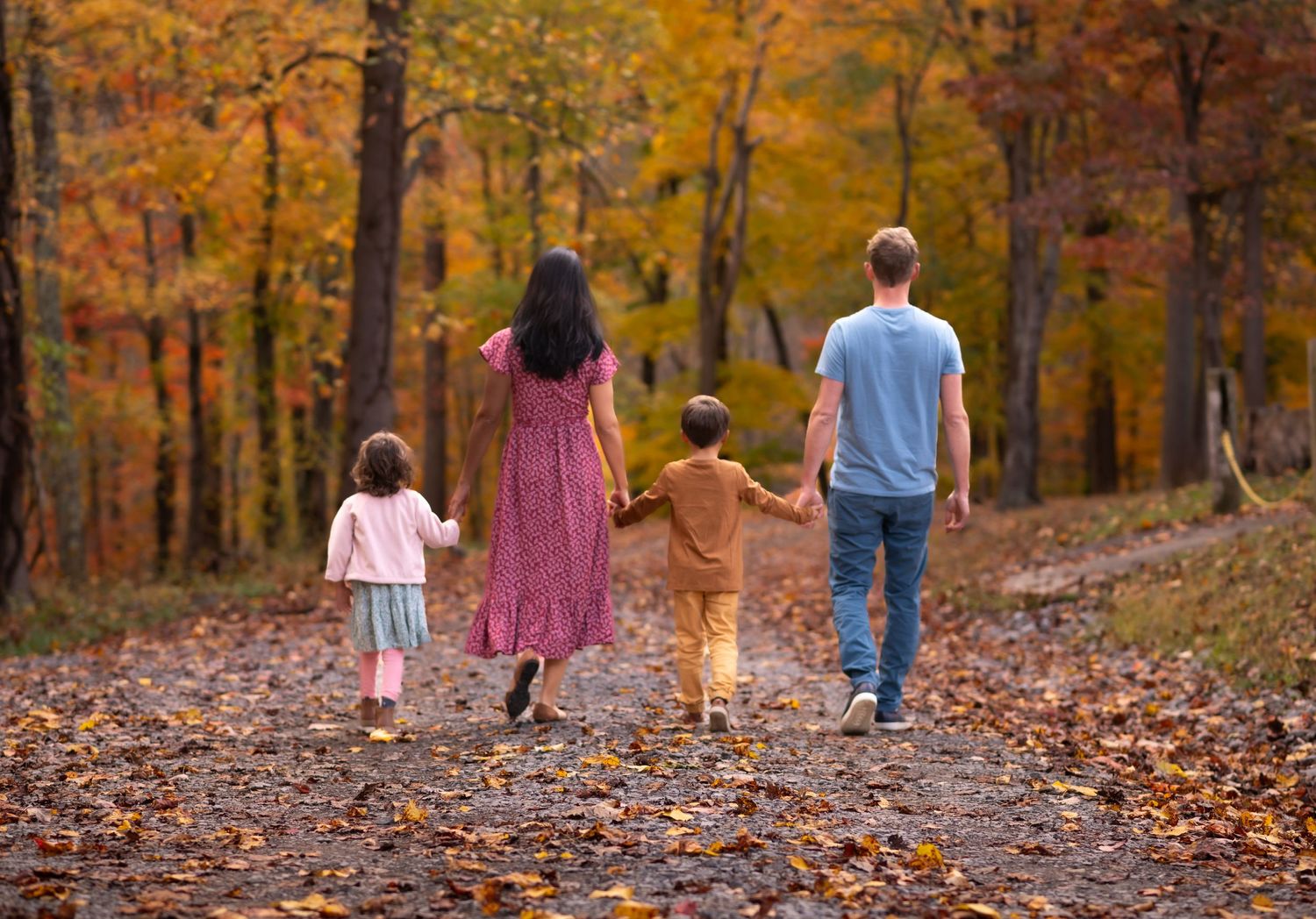 A father, mother, and two young children in coordinating outfits walk away from the camera down a gravel road with fall leaves all around.