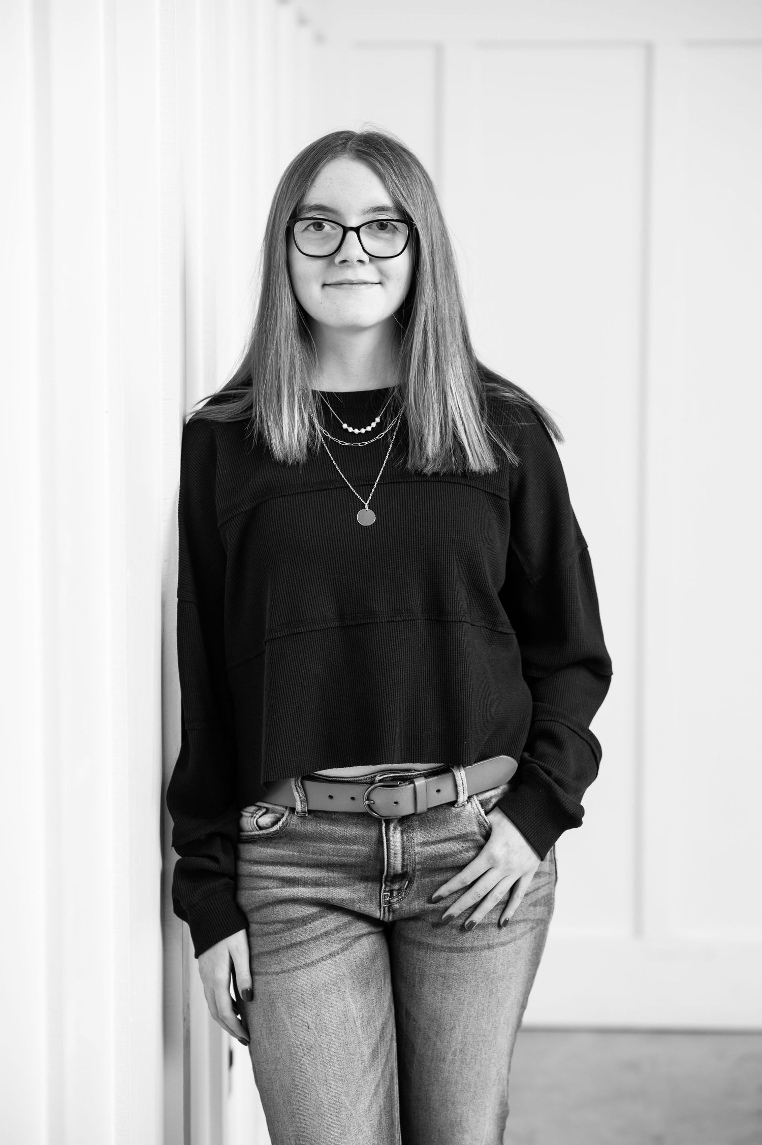 black and white casual senior portrait of a young woman with straight brown hair and glasses and a black sweater, leaning against a wall