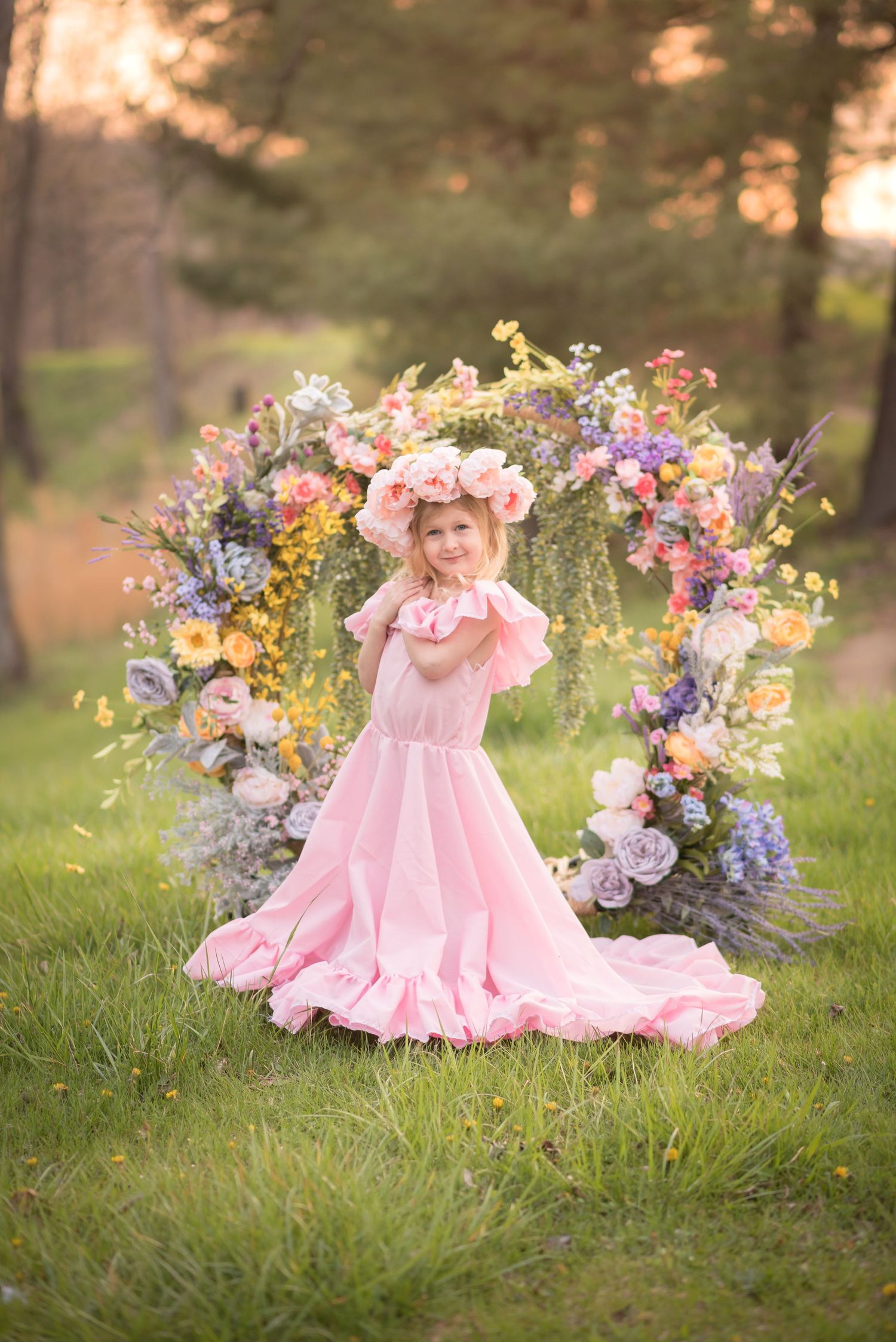 a little girl in a long ruffled pink dress and flower crown poses in front of a floral arch