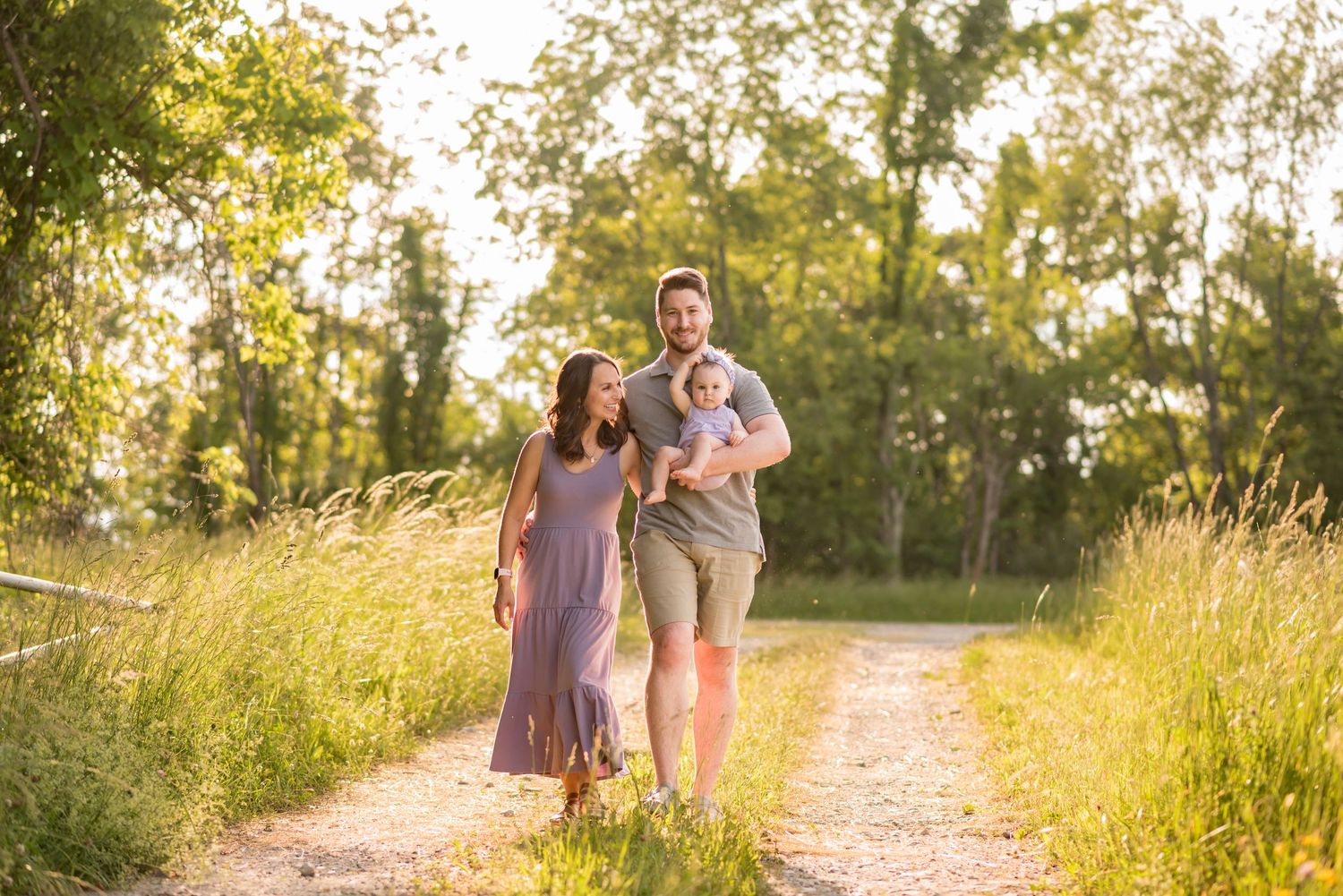 a young couple stroll down a sunlit farm path hand in hand, with the husband holding their baby girl in his arm; trees and field behind them
