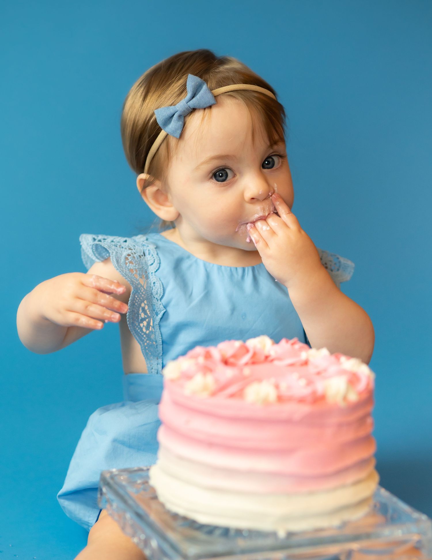 a baby girl in a blue dress sits on a blue studio backdrop with a pink and white birthday smash cake, with a handful of icing in her mouth