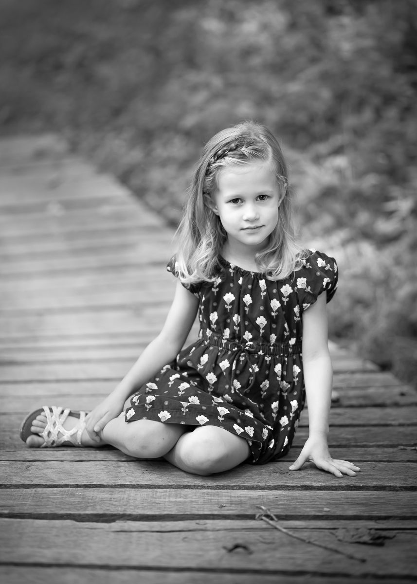 a little girl in a blue flowered dress sits on a wooden bridge and looks solemnly at the camera