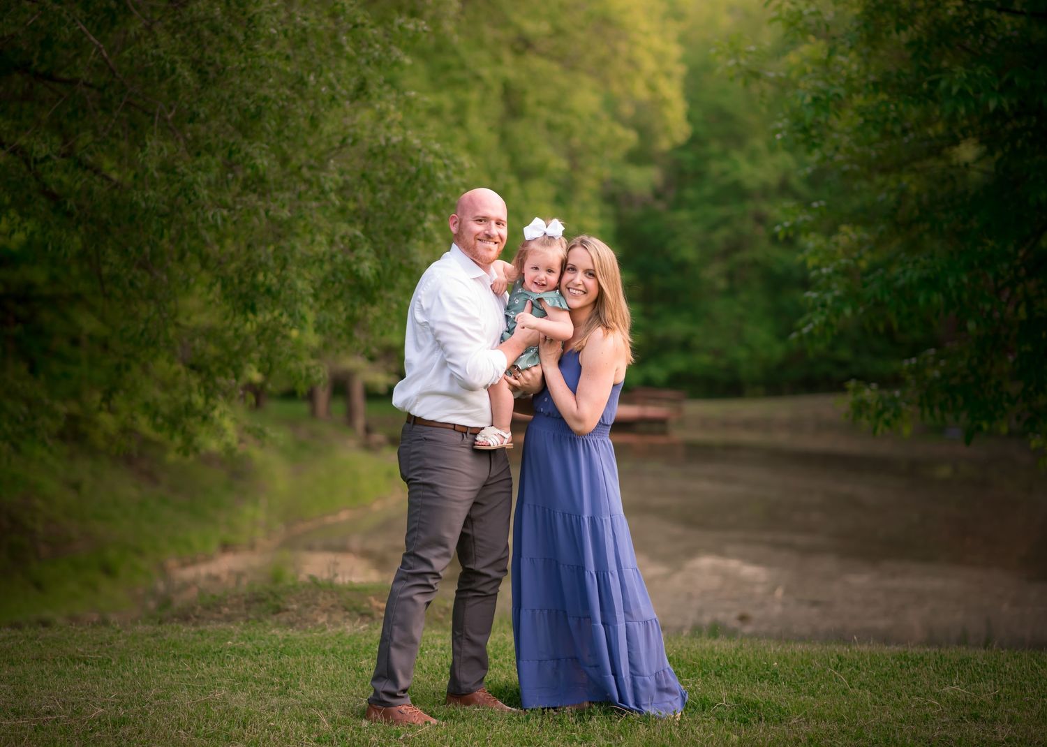 a couple and their toddler daughter stand together in the grass and pose for a portrait, smiling at the camera with trees all around