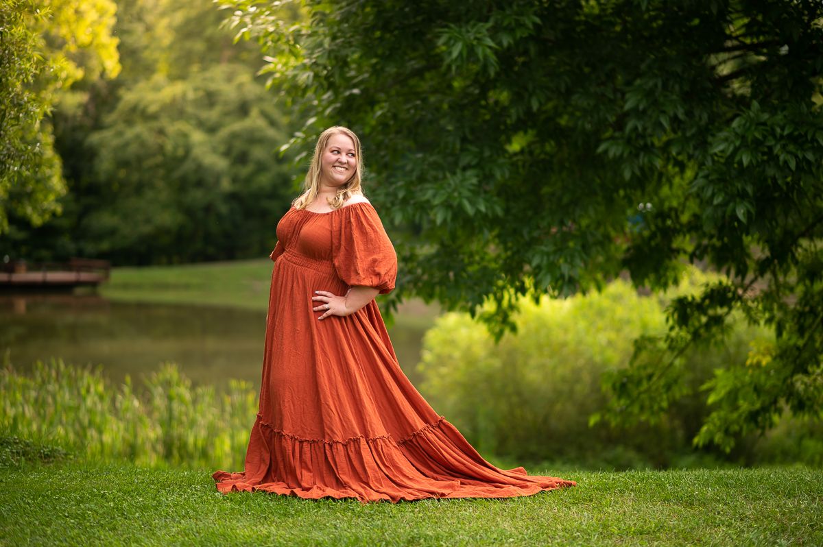 a lovely young blonde woman in a long orange dress poses with a smile in front of a pond, surrounded by lush green trees and grass