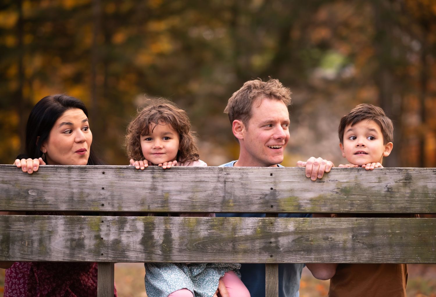 a family of four peek over the edge of a wooden bench and make silly faces for a fun portrait