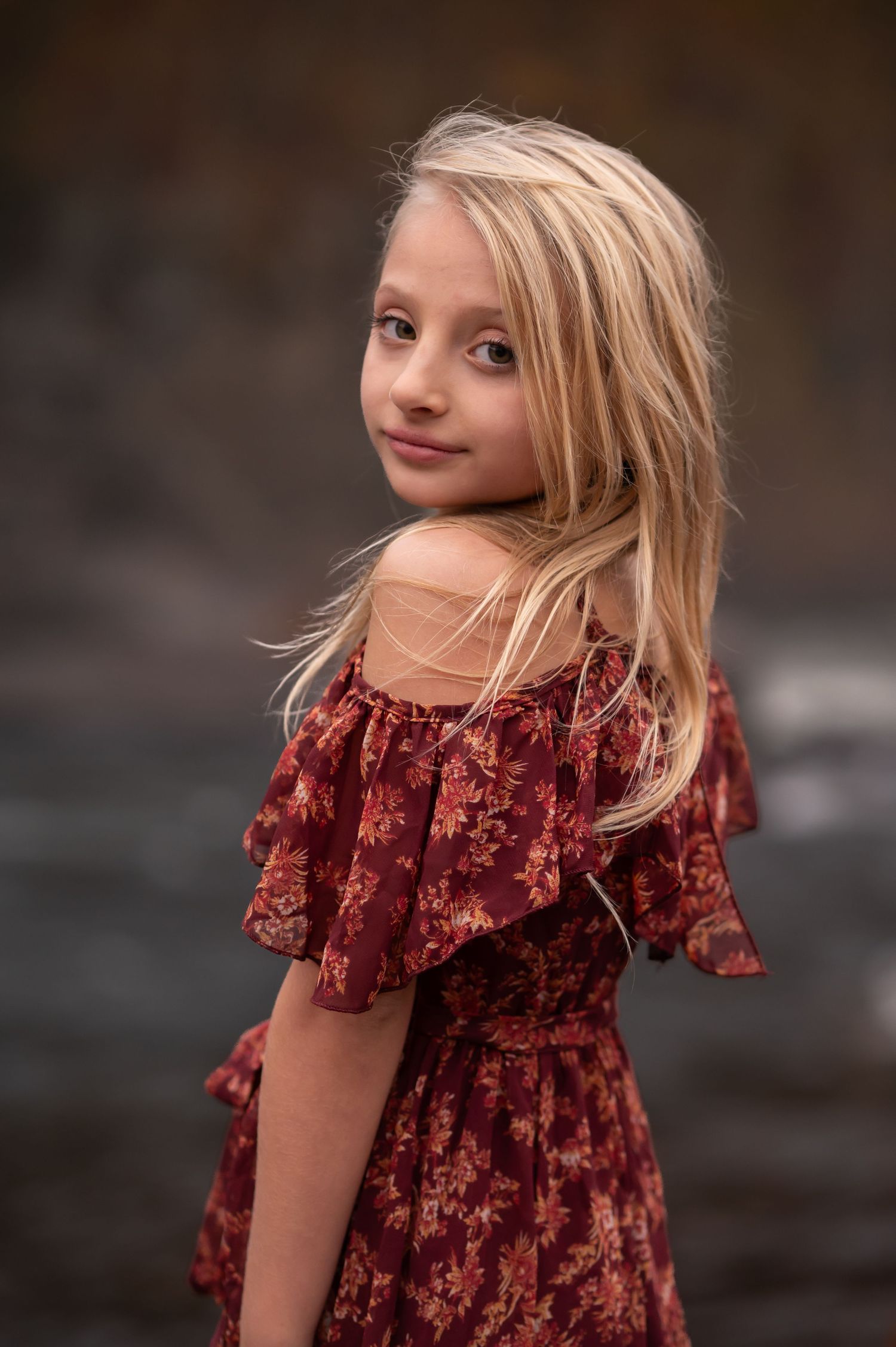 closeup portrait of a young blonde girl in an off the shoulder red floral dress, standing in front of a waterfall at Valley Falls State Park