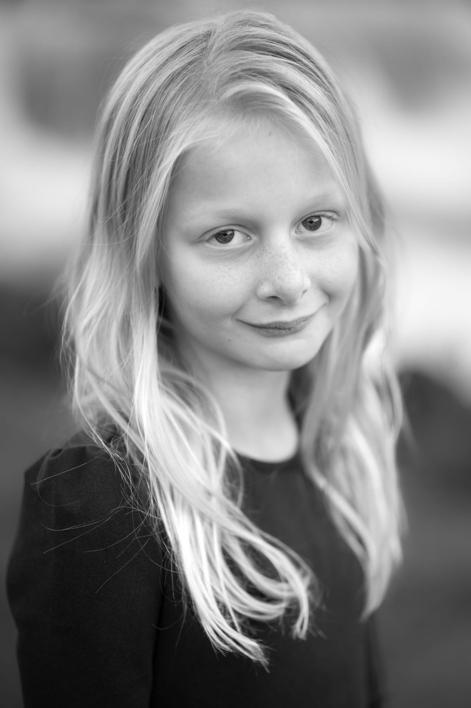 black and white closeup portrait of a little girl with long blonde hair and freckles across her nose, smiling shyly at the camera