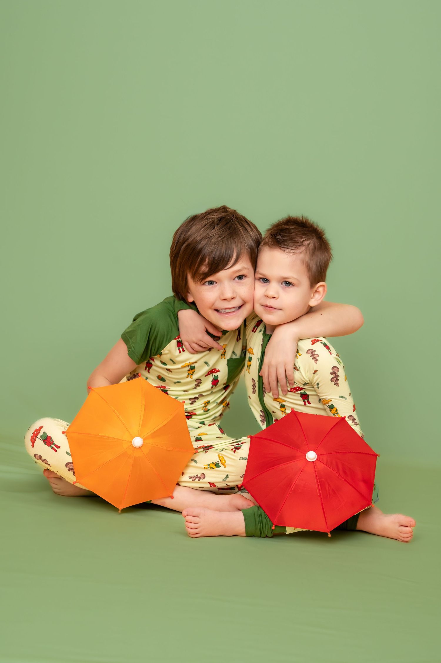 two little boys in matching green pajamas pose together on a green studio background with brightly colored orange and red umbrellas