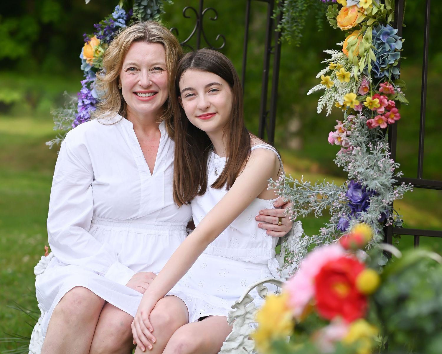 a mother and daughter in matching white dresses sit together on a white bench under and arch of flowers