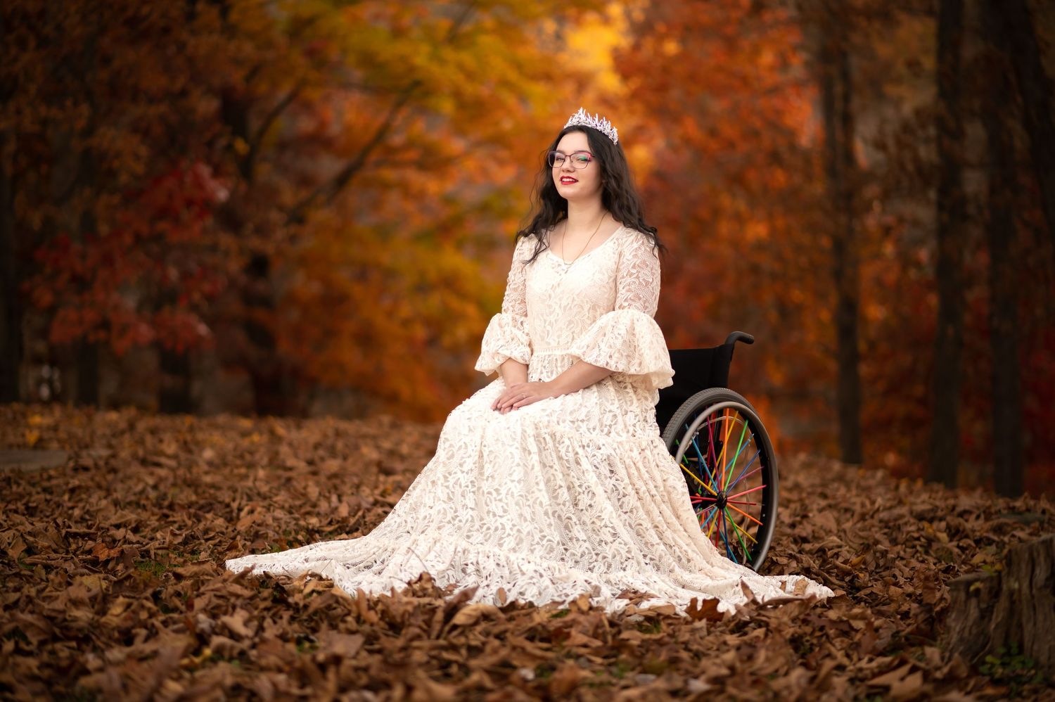 a young woman in a long white lace dress sits in a wheelchair surrounded by fallen autumn leaves, with brilliant fall foliage in background