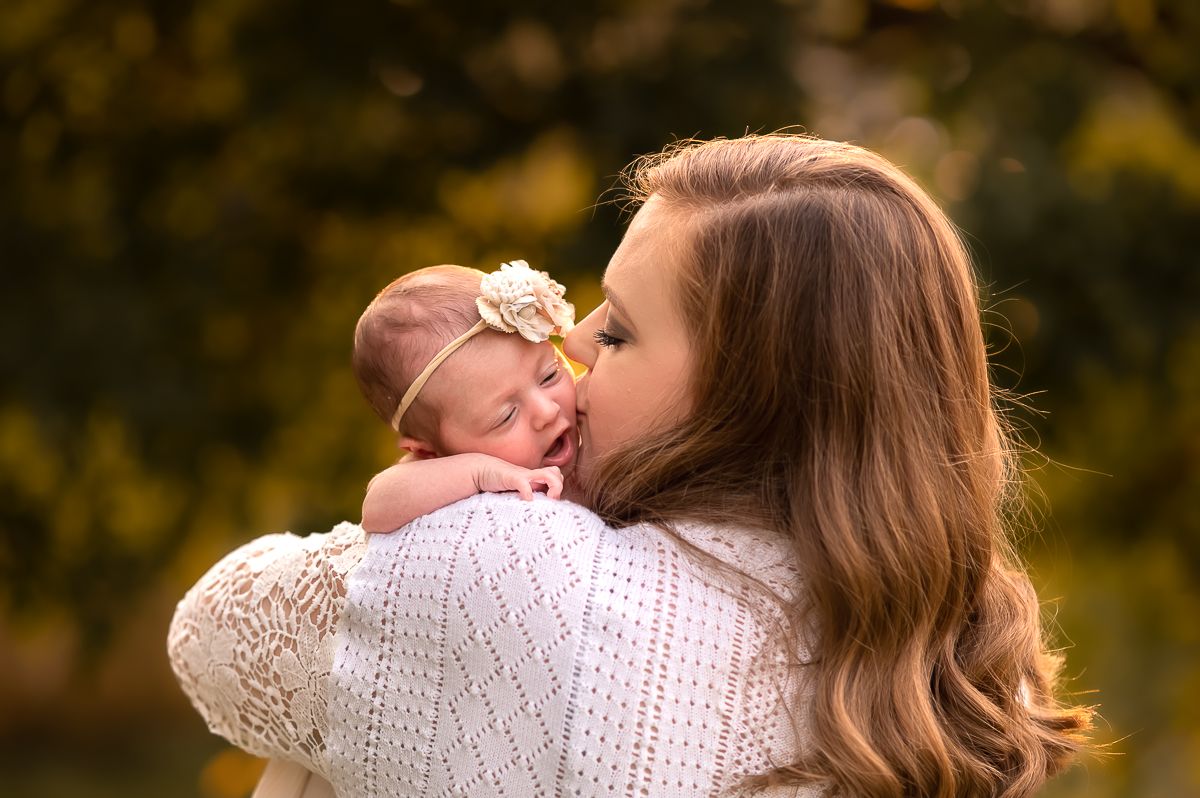 a young mother in a white sweater holds her newborn baby up to her shoulder and kisses her cheek; taken outside in the fall in Morgantown