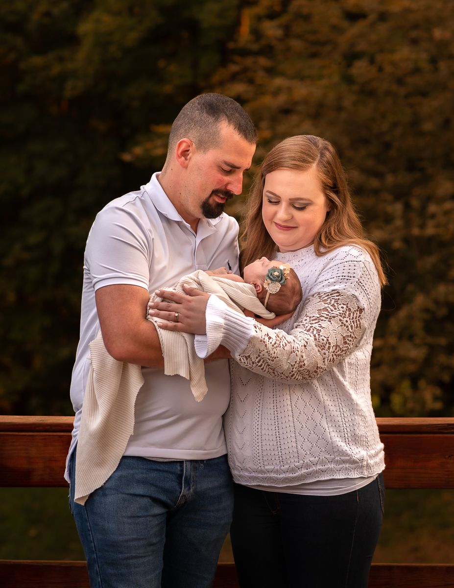 A young couple hold their infant daughter and smile at her, while standing on a wooden bridge with fall scenery all around in Morgantown