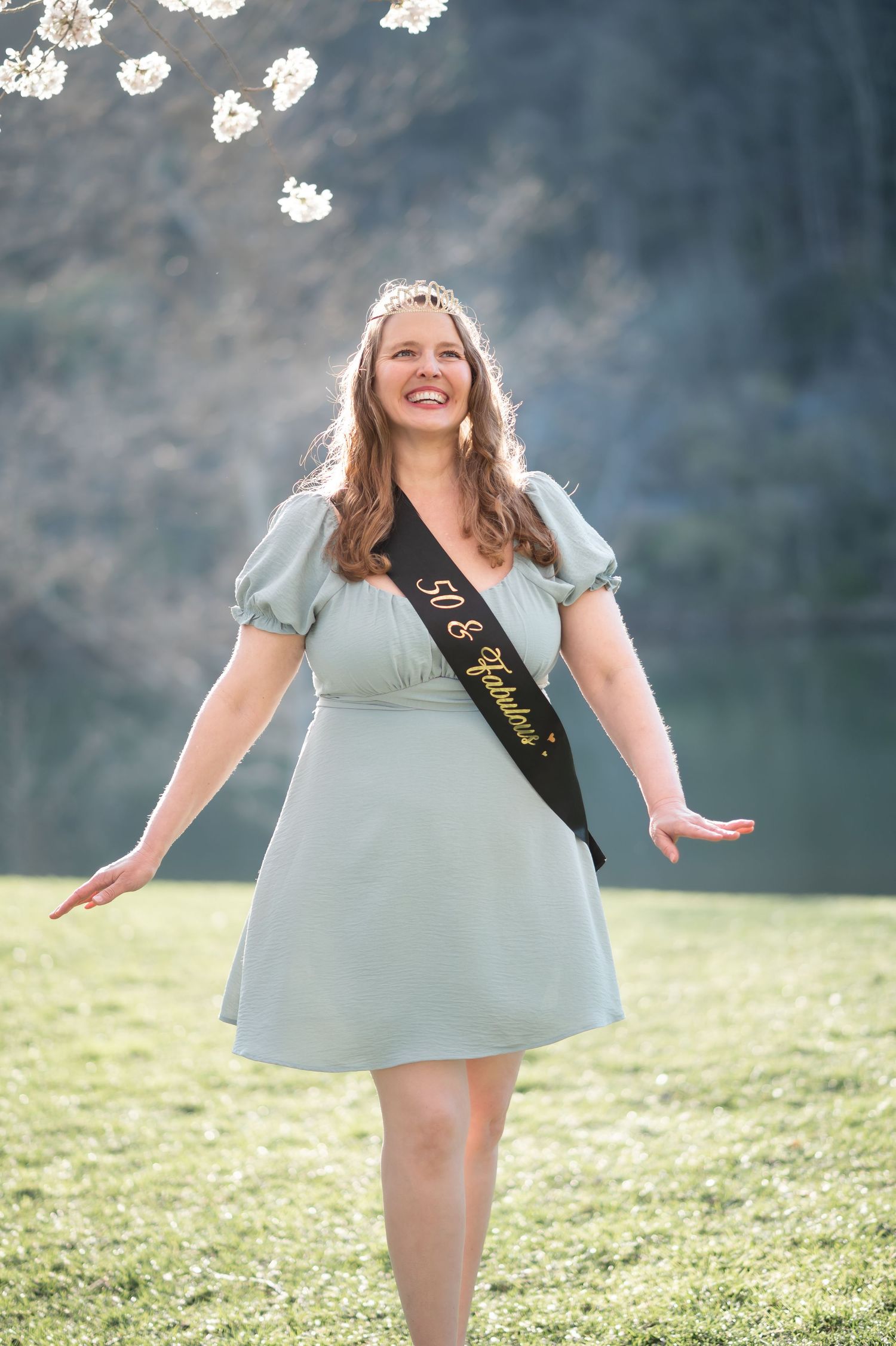 a woman with long brown hair and a light blue dress, crown, and 50th birthday sash laughs and twirls under cherry blossoms in Morgantown, WV