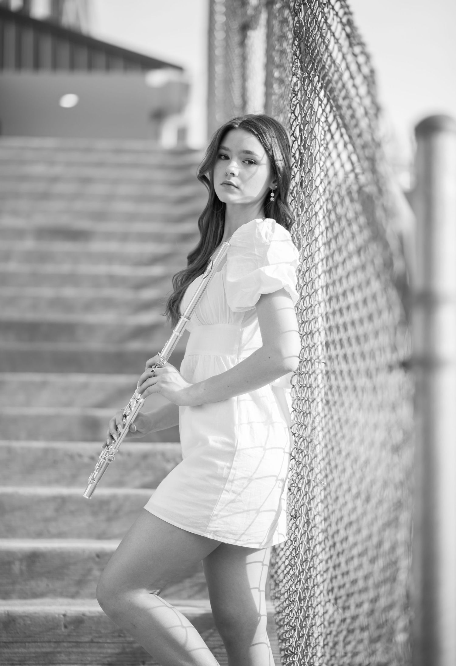 black and white senior portrait of a young woman in a white dress, standing by a chainlink fence in front of concrete stairs holding a flute