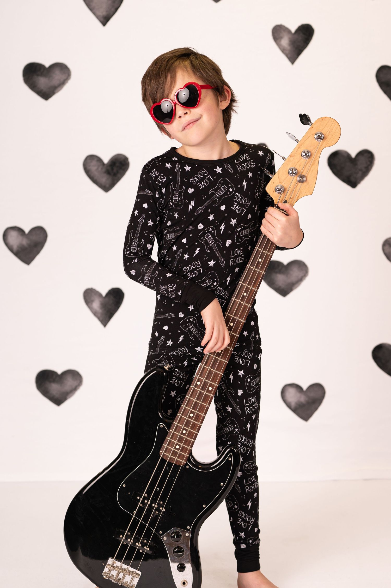 a little boy in red heart shaped sunglasses and black pajamas poses with an electric guitar with a white and black studio background