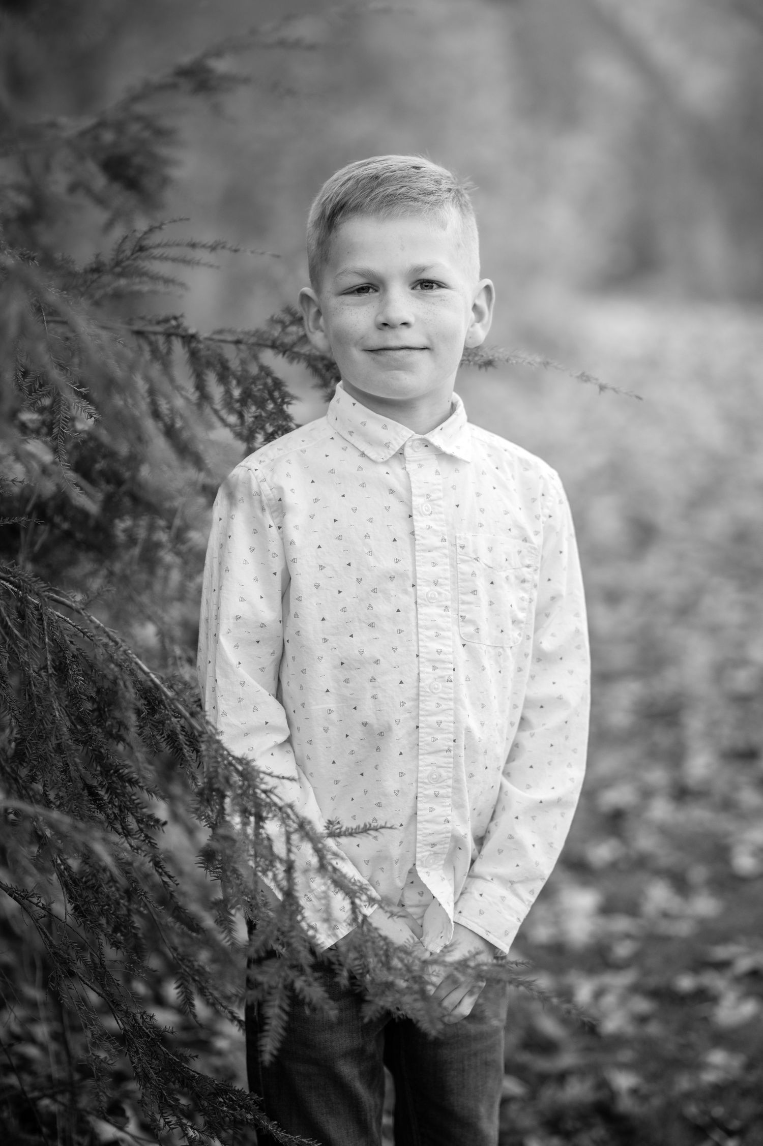 black and white; a young boy in a white button up shirt poses for a portrait outside beside a large evergreen tree