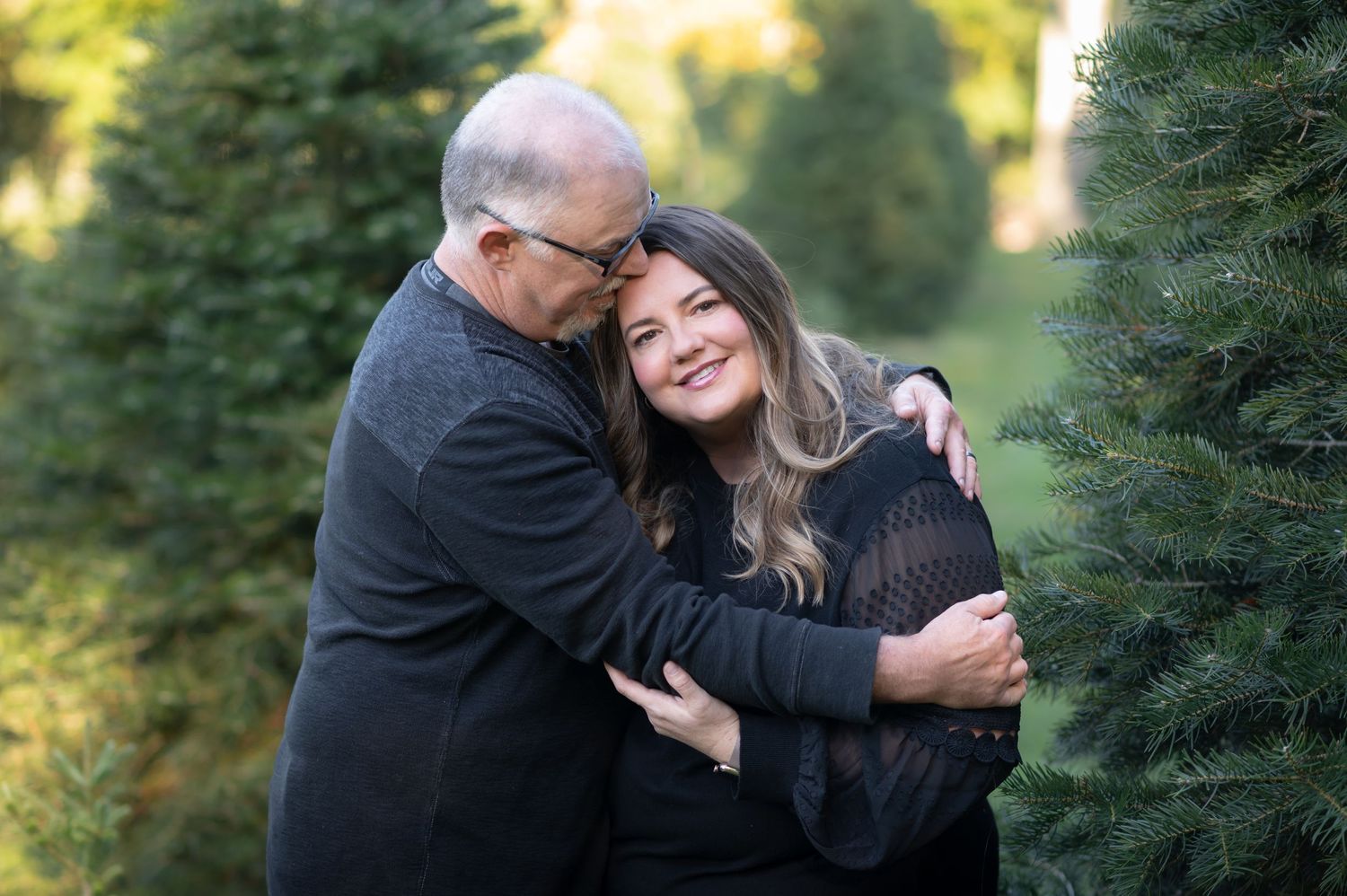 a man and woman pose for a portrait in the middle of a Christmas tree farm; he hugs her while she smiles at the camera.