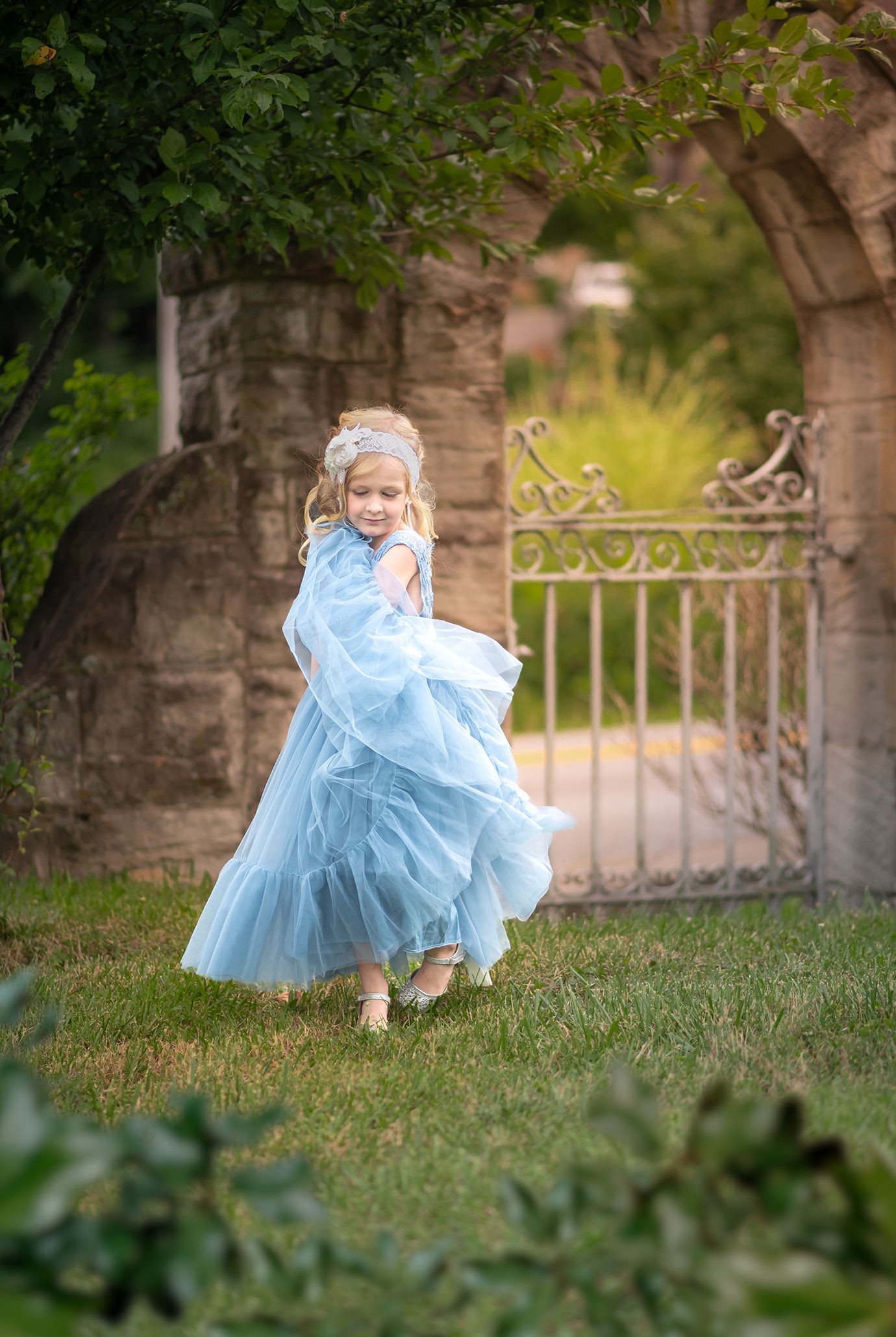a little girl in a fancy blue tulle dress and a floral headband twirls in front of an old stone gate
