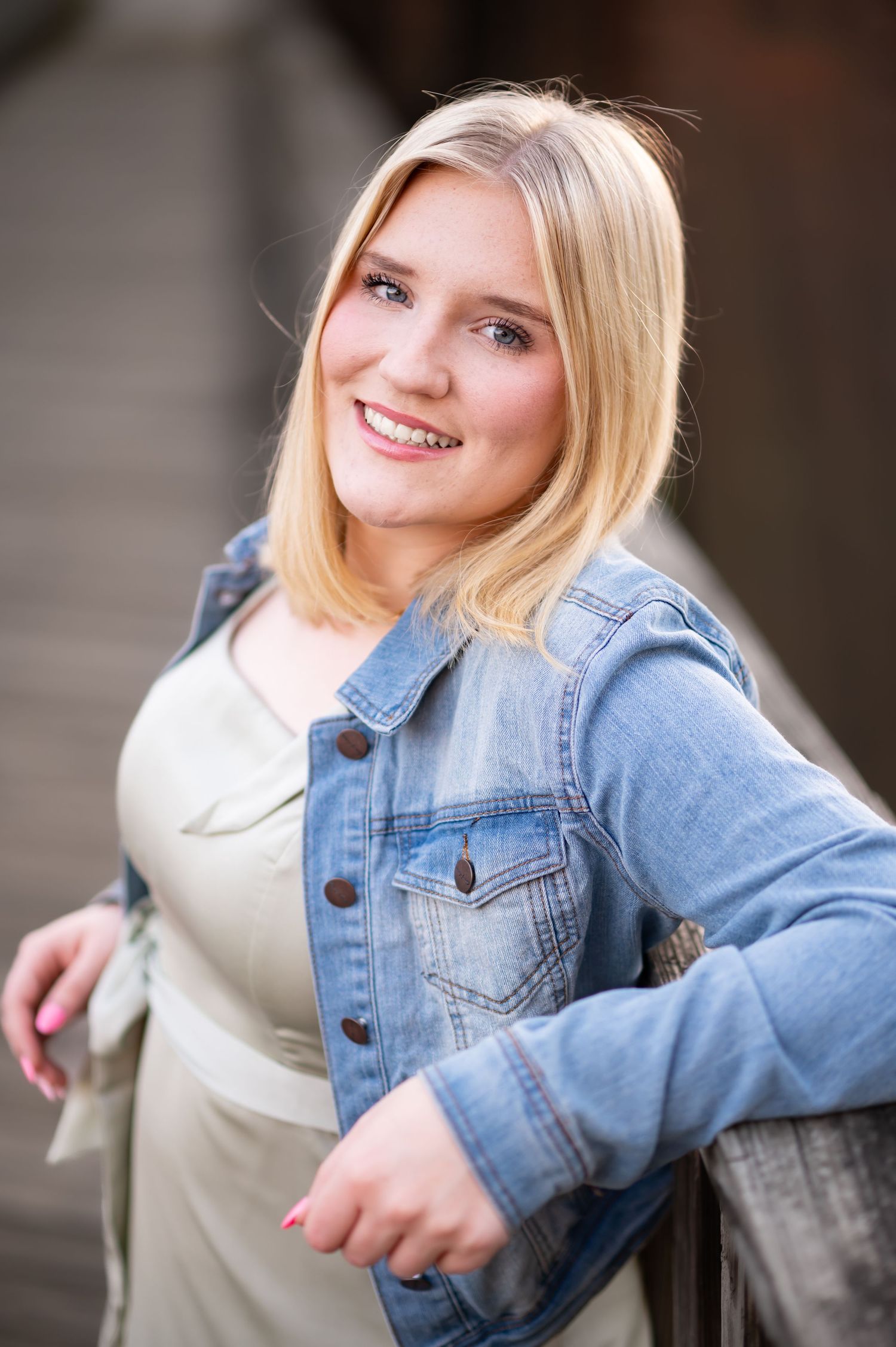 a blonde high school graduate in a denim jacket leans against a wood bridge and smiles into the camera for a closeup senior portrait