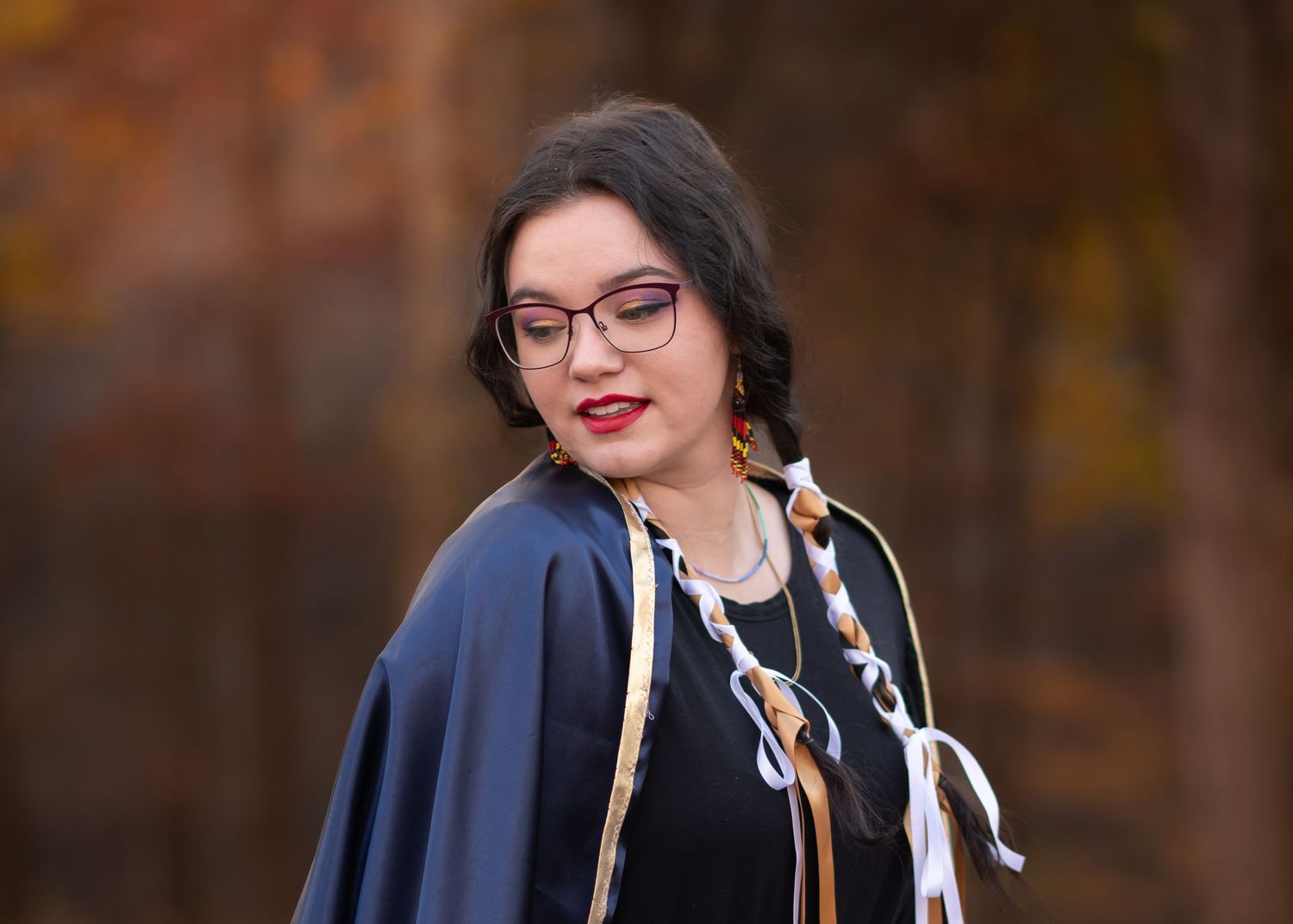 portrait of a young woman with long dark braided hair wearing a traditional indigenous American ceremonial outfit with fall leaves behind