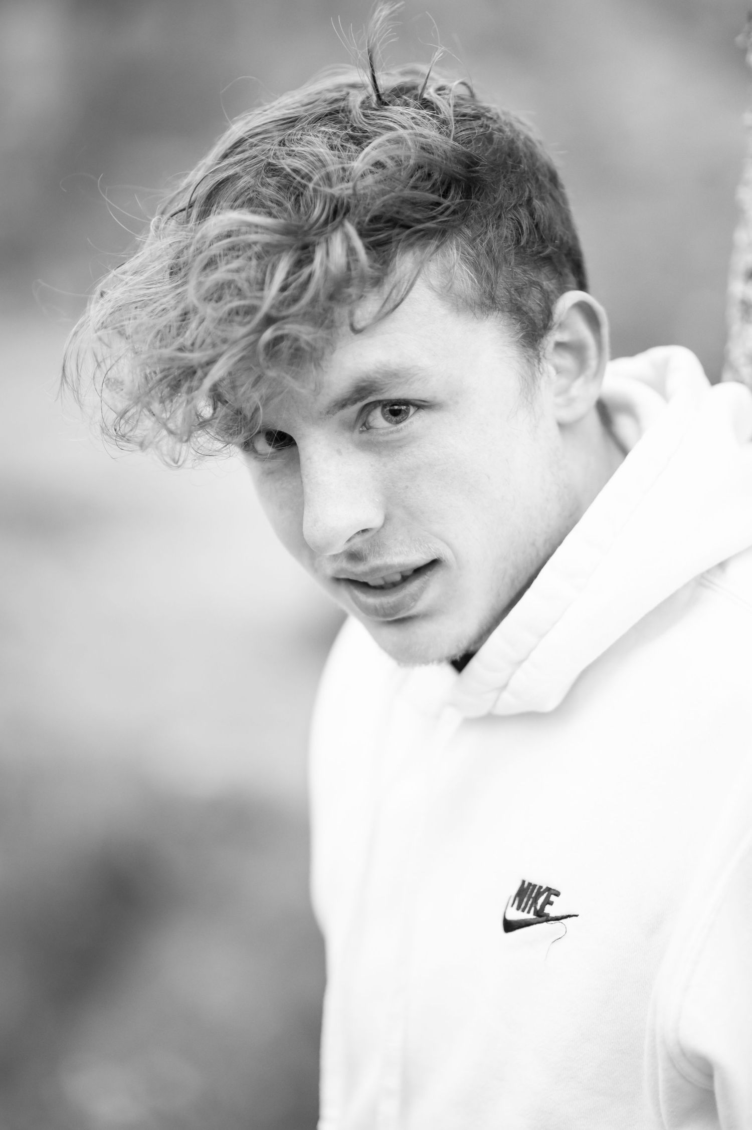 closeup black and white portrait of a high school boy with curly hair in a white hoodie, leaning against a tree with a river behind him