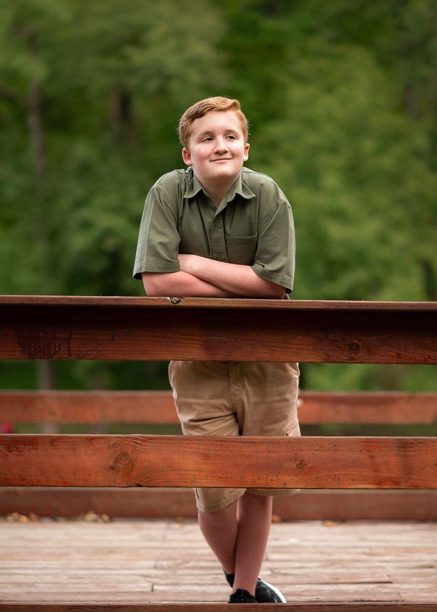 a young man in a green polo shirt leans his elbows on a wooden railing and smiles for the camera