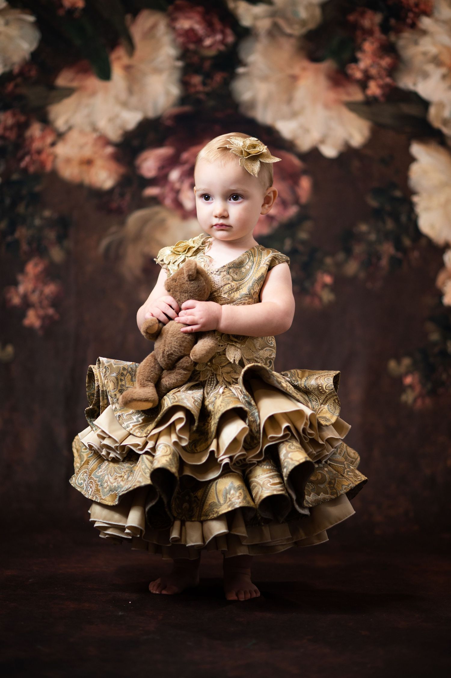 a little girl in a ruffled gold brocade couture dress stands in front of a classical floral studio background for a portrait