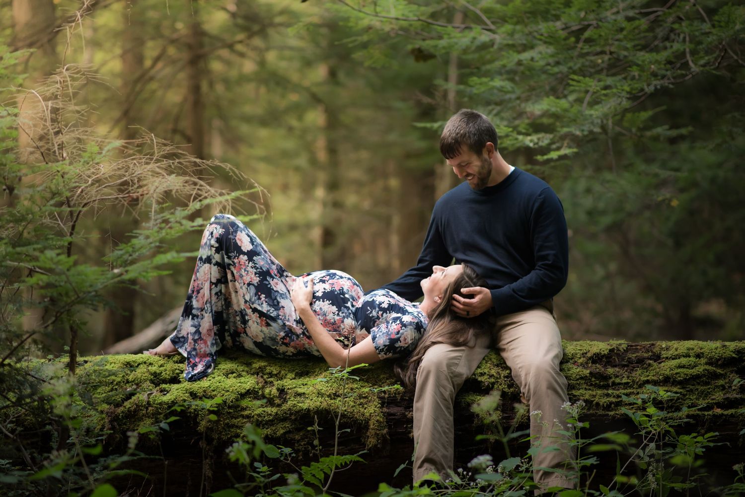 a young pregnant woman in a long floral dress and her husband sit on a mossy fallen log in the middle of the woods with sunlight all around