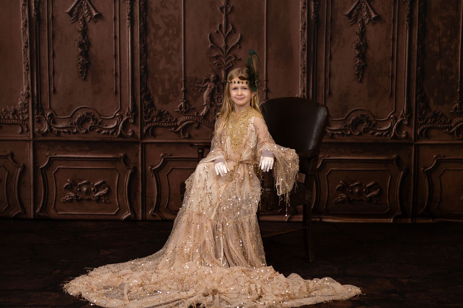 dramatic classic portrait of a little girl in a long gold sequin dress, gold gloves and a feathered headdress, seated in a leather chair