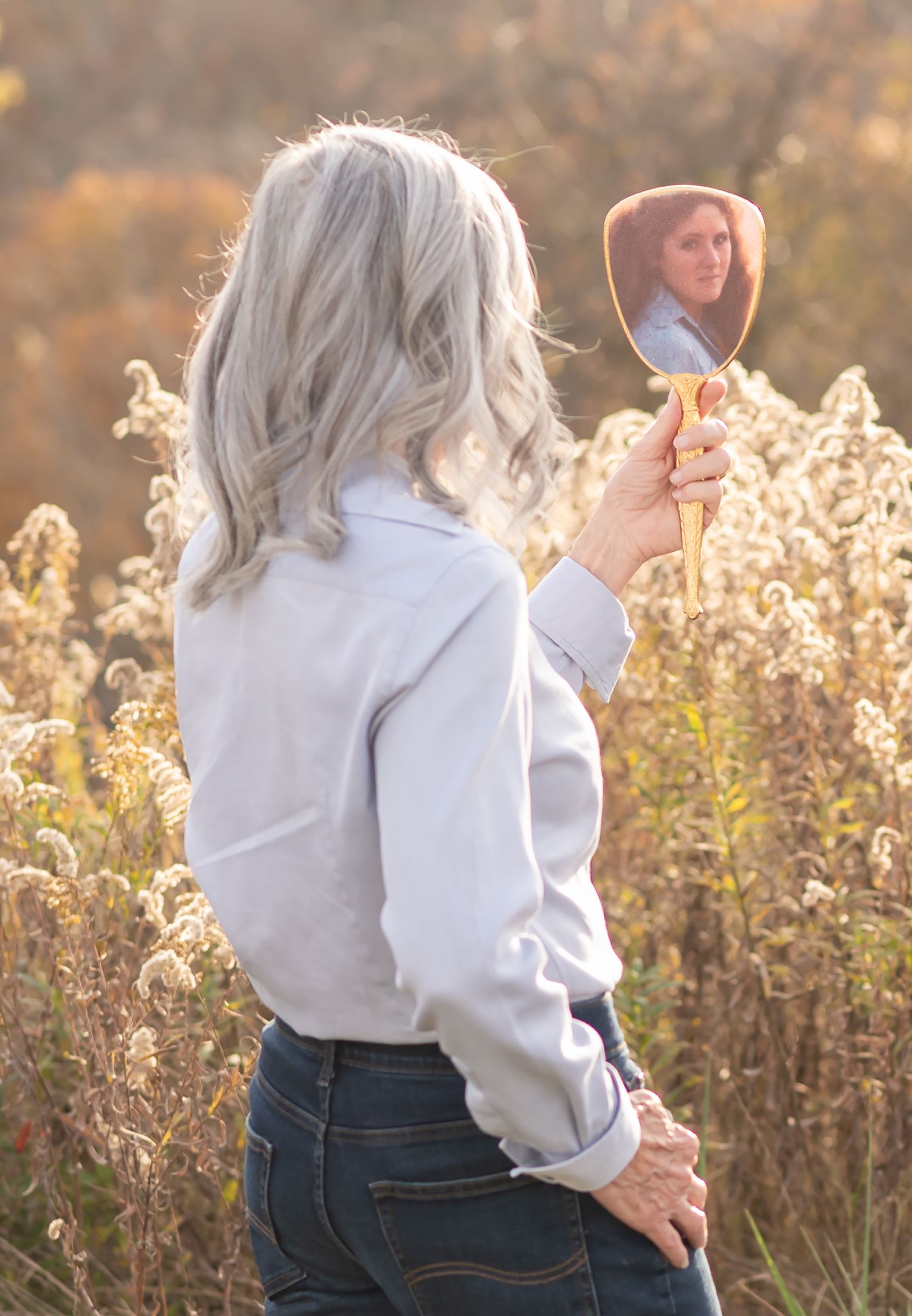 an older woman in a light blue top standing in a wheat field holding a hand mirror and gazing at the reflection of her younger self