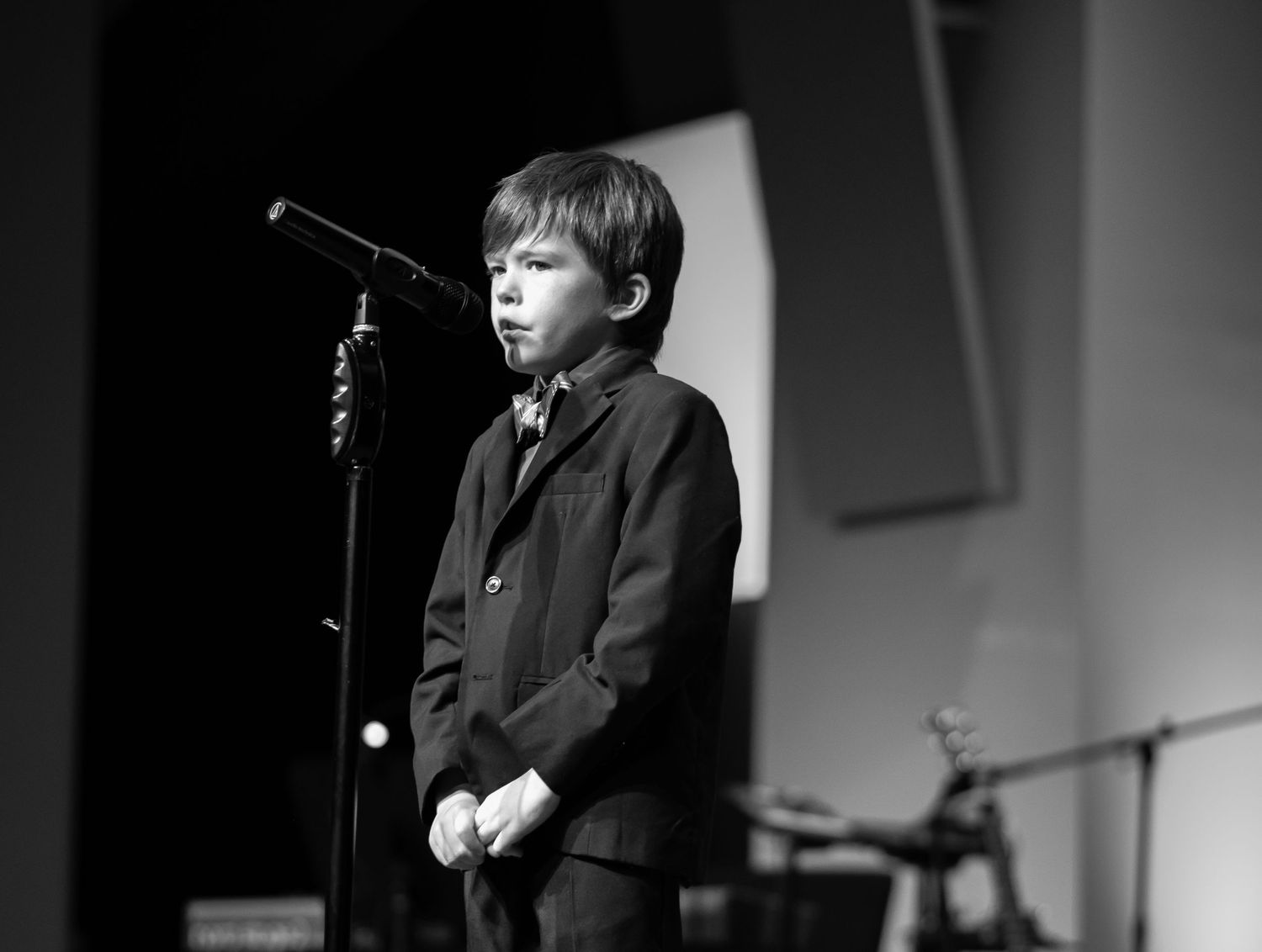 black and white event portrait of a little boy in a suit and tie, standing nervously at a microphone on a large stage