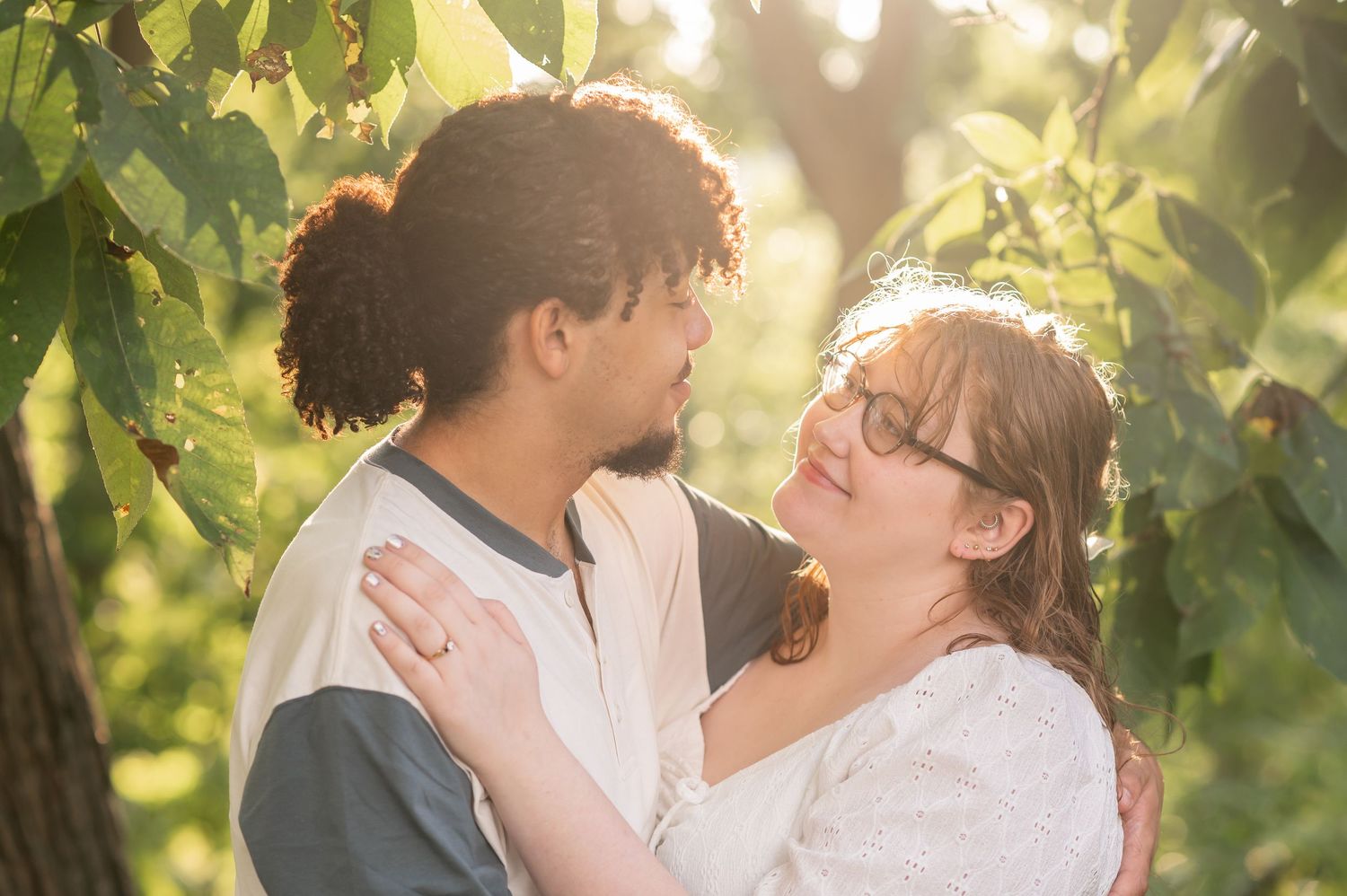 a young engaged couple embraces and smiles at each other, surrounded by sunlit trees in Morgantown, WV