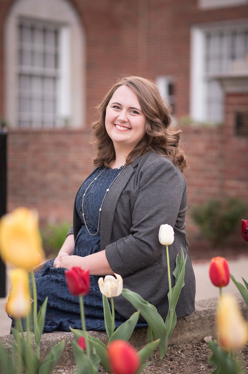 a young graduate student in a navy top and gray blazer poses for a graduation portrait in front of Elizabeth Moore Hall, with tulips