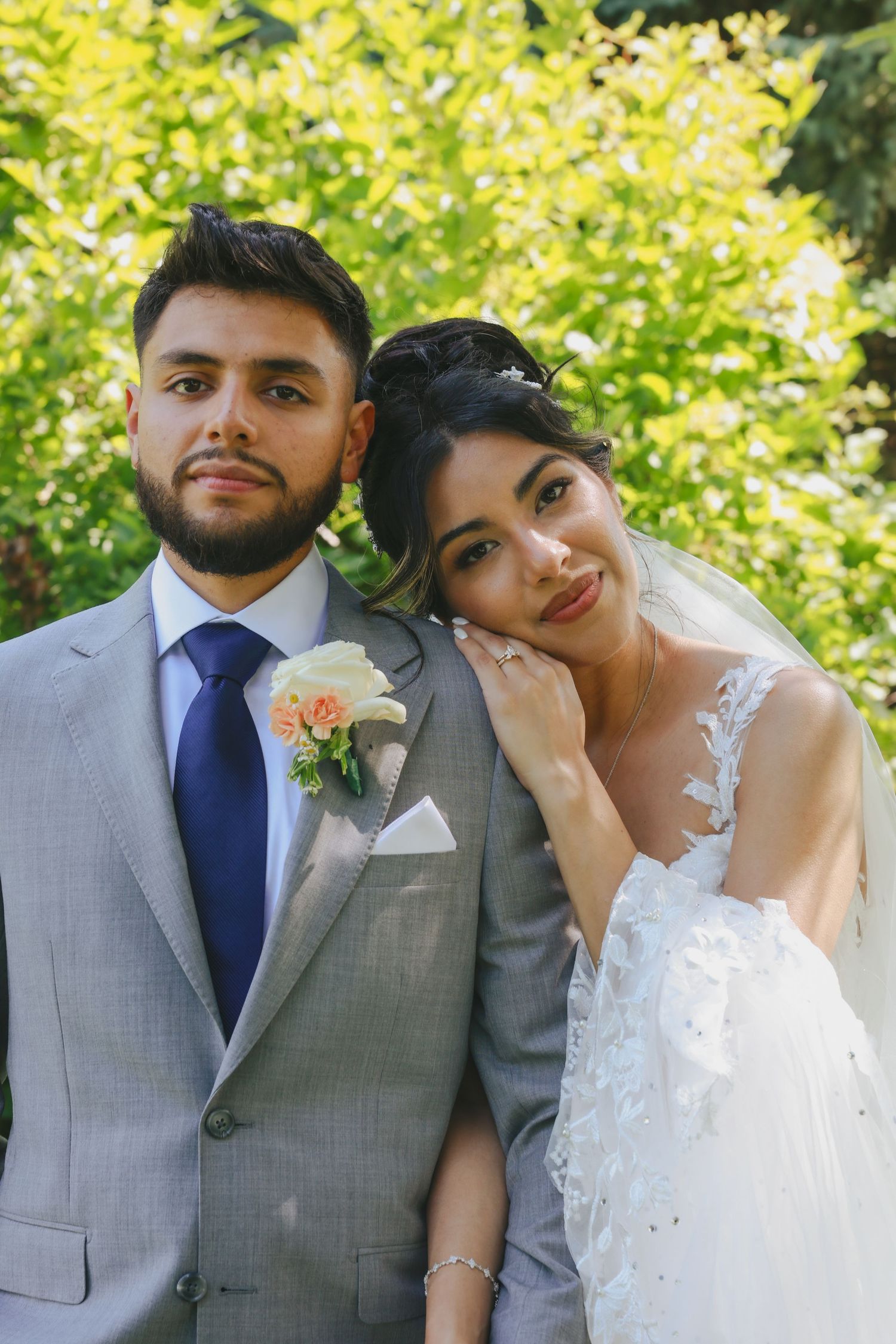 A wedding couple in traditional formal attire pose together outdoors against green foliage on their special day.